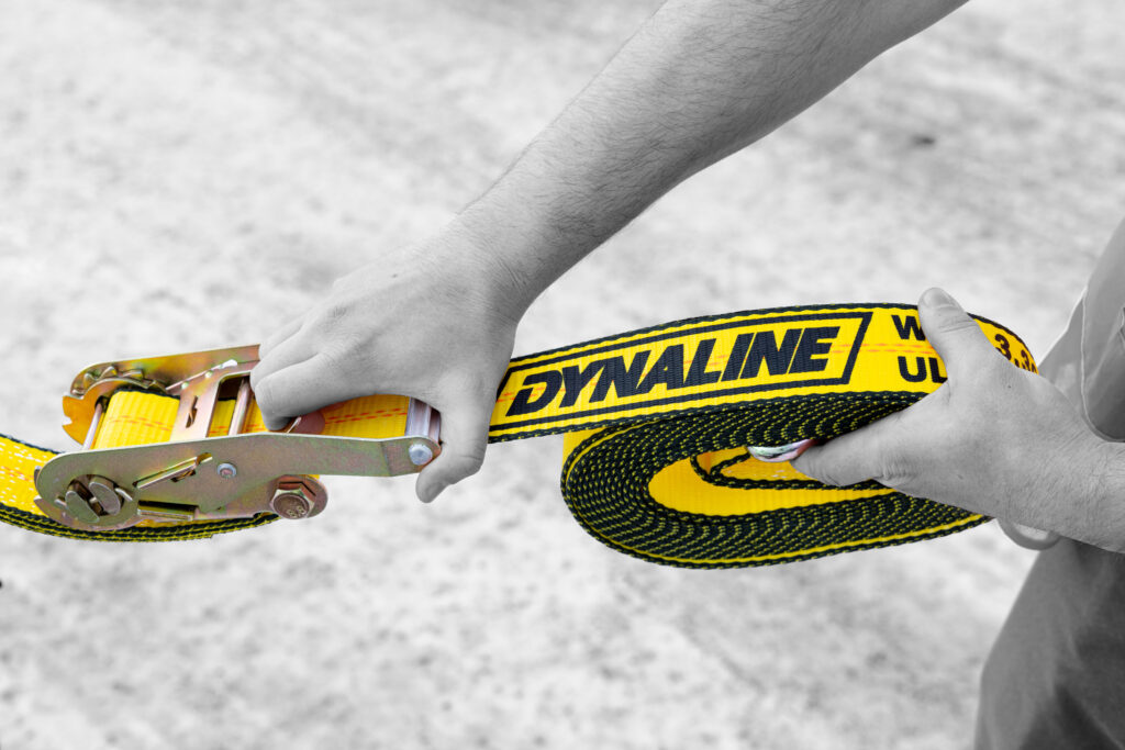 Hand pulling a yellow Dynaline ratchet strap across the ground